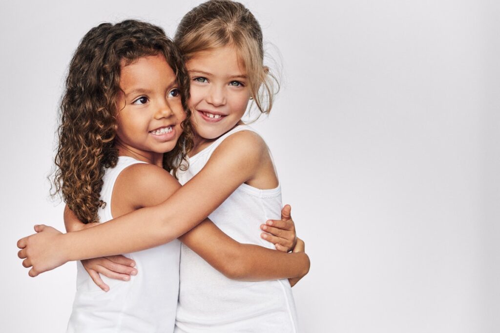 Zwei kleine Mädchen umarmen sich. Sie tragen weiße Unterhemden.