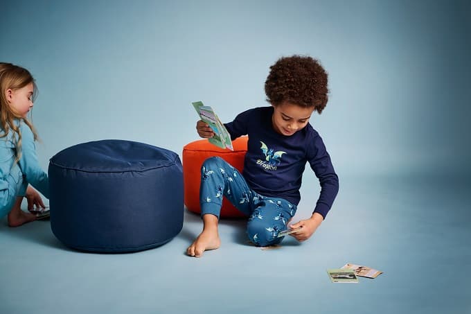 Zwei Kinder spielen auf dem Boden in gemütlicher Umgebung. Der Junge in einem dunkelblauen Pyjama mit leuchtendem Drachenmotiv sitzt auf einem orangen Sitzhocker und hält Spielkarten in der Hand. Ein Mädchen in hellblauer Nachtwäsche kniet im Hintergrund neben einem dunkelblauen Pouf.