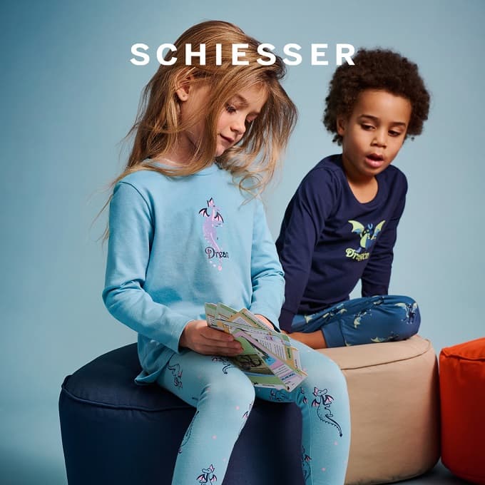 Zwei Kinder in gemütlicher Nachtwäsche von SCHIESSER sitzen auf Sitzhockern vor blauem Hintergrund; das Mädchen trägt einen hellblauen Pyjama mit Drachenmotiv und hält Karten in der Hand, der Junge trägt einen dunkelblauen Pyjama mit leuchtendem Drachenaufdruck.