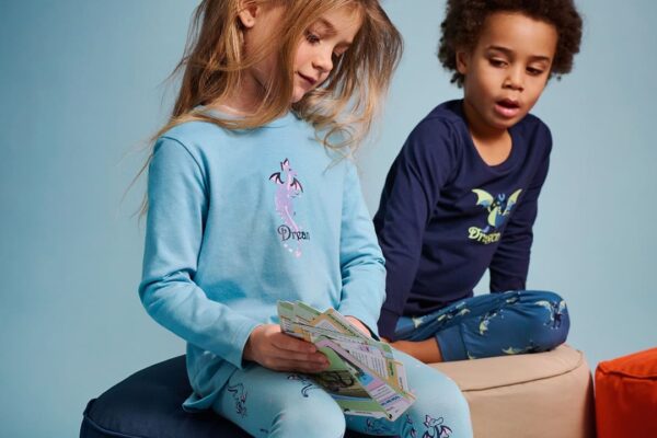 Zwei Kinder in gemütlichen Glow in the Dark Pyjamas von SCHIESSER sitzen auf Sitzhockern vor blauem Hintergrund; das Mädchen trägt einen hellblauen Pyjama mit Drachenmotiv und hält Karten in der Hand, der Junge trägt einen dunkelblauen Pyjama mit leuchtendem Drachenaufdruck.