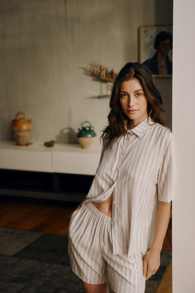 Frau in lockerer, gestreifter Nachtwäsche lehnt entspannt an einer Wand – bequemer Schnitt für optimalen Komfort in Übergrößen und Sonderlängen für Damen.