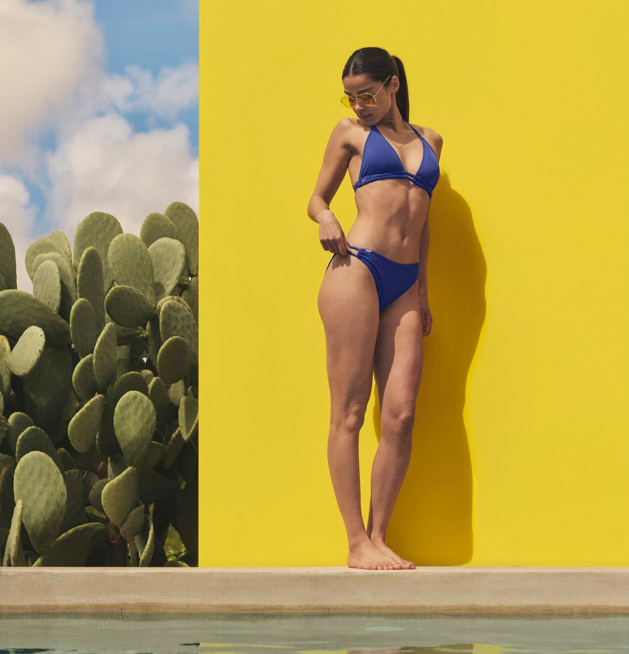 Frau trägt einen blauen Bikini am Pool vor einer gelben Wand.
