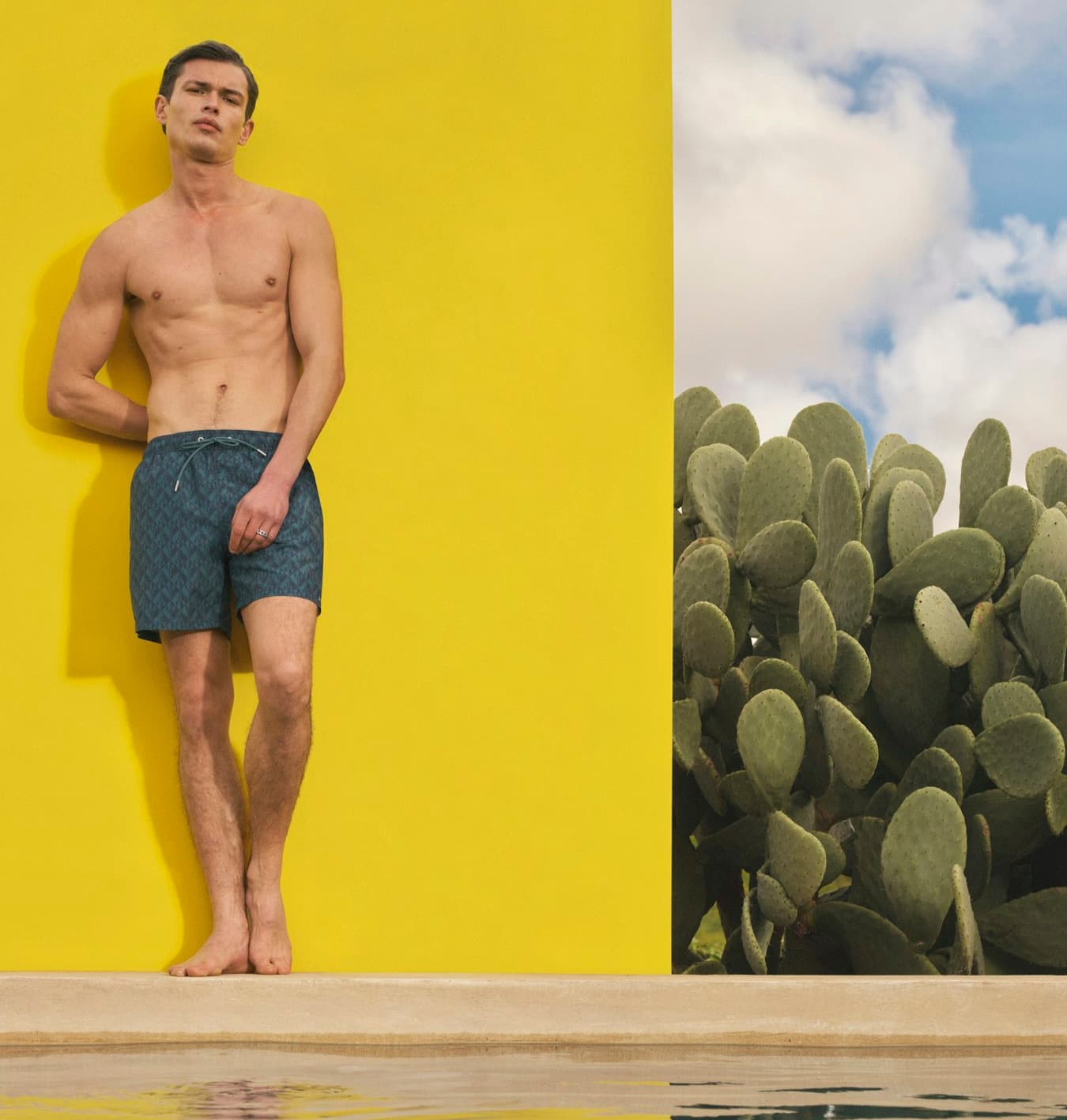 Mann trägt eine dunkelblaue Badehose am Pool vor einer gelben Wand.
